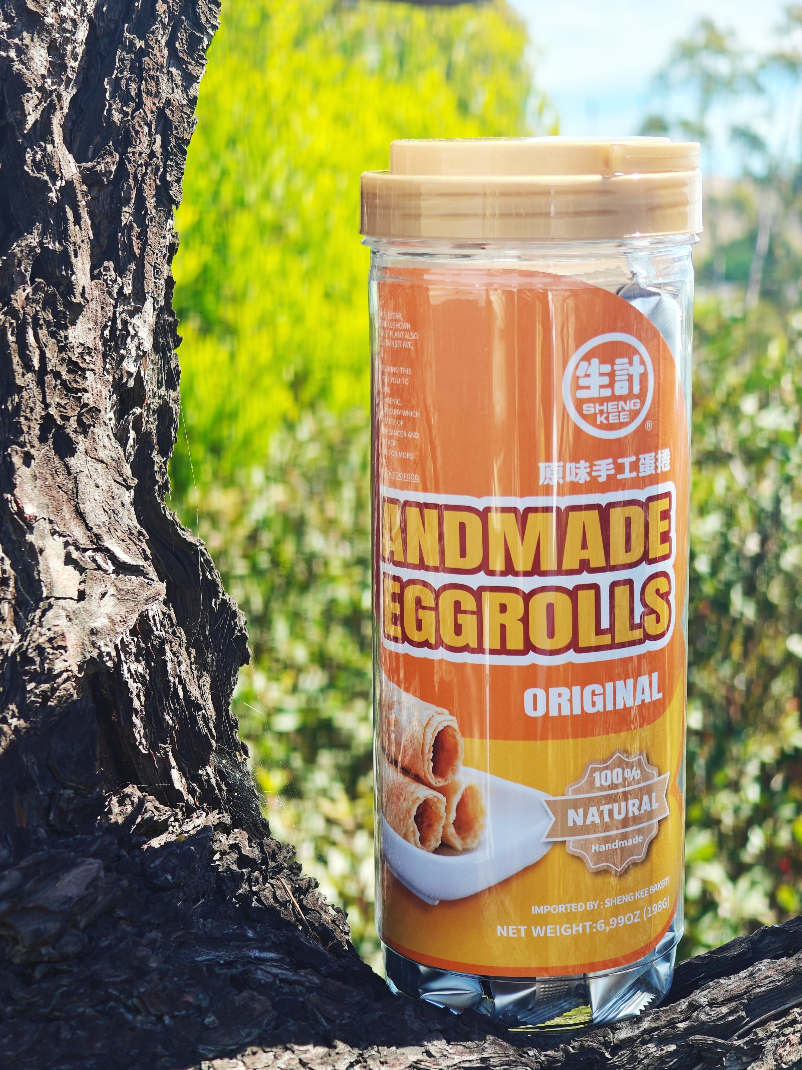 A clear plastic jar of Handmade Eggrolls with a beige lid and orange label, set on a tree trunk outdoors with a blurred green background.