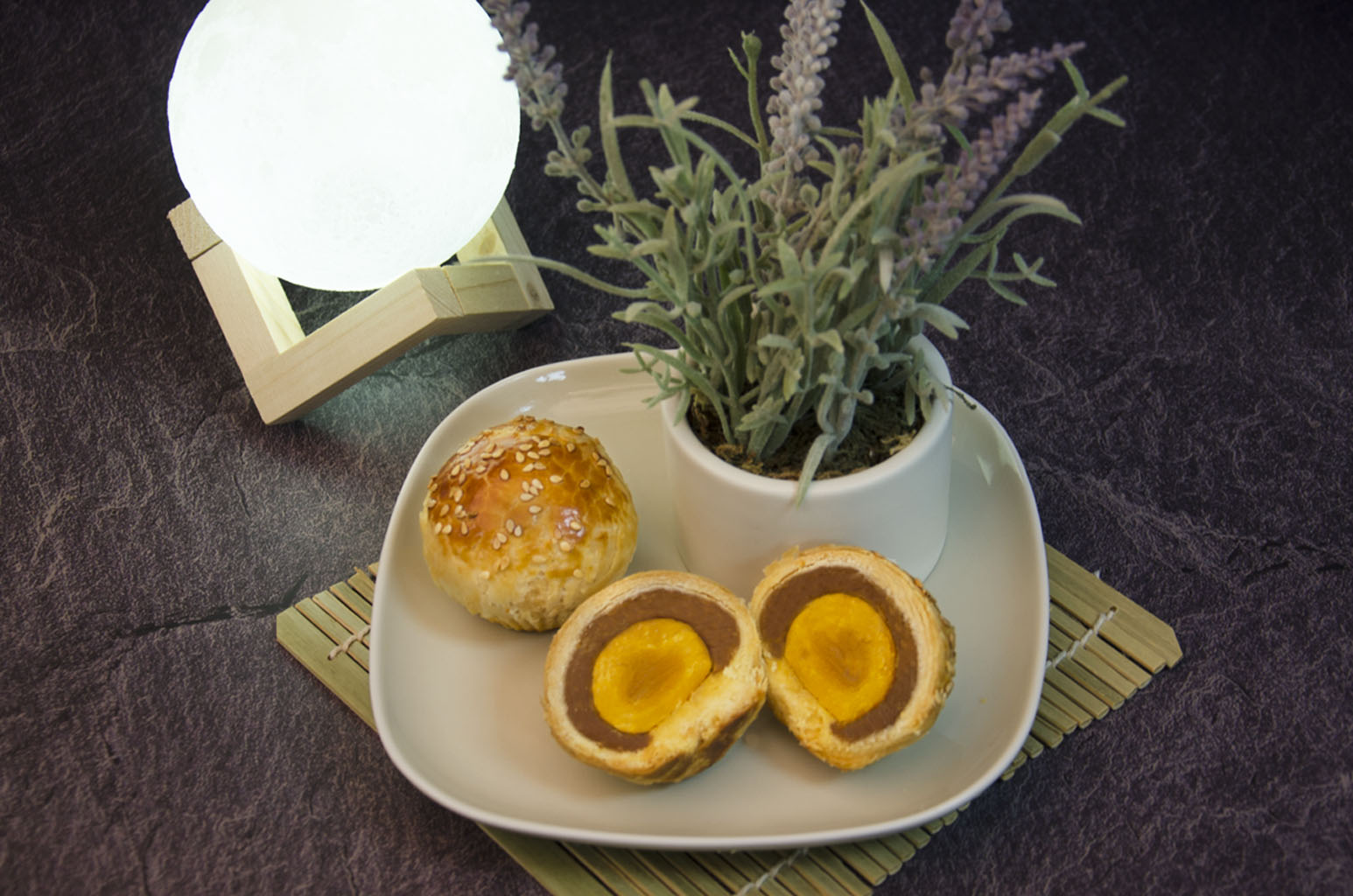 Lotus Salted Egg Yolk Pastry. One cut open to see cross-section on a white plate with small plant in vase also on plate. In front of white ball of light on wooden contraption.
