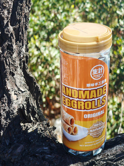 A jar of Handmade Eggrolls placed on a tree trunk outdoors with a blurred green forest background. The jar has a yellow lid and an orange label with white and yellow text.