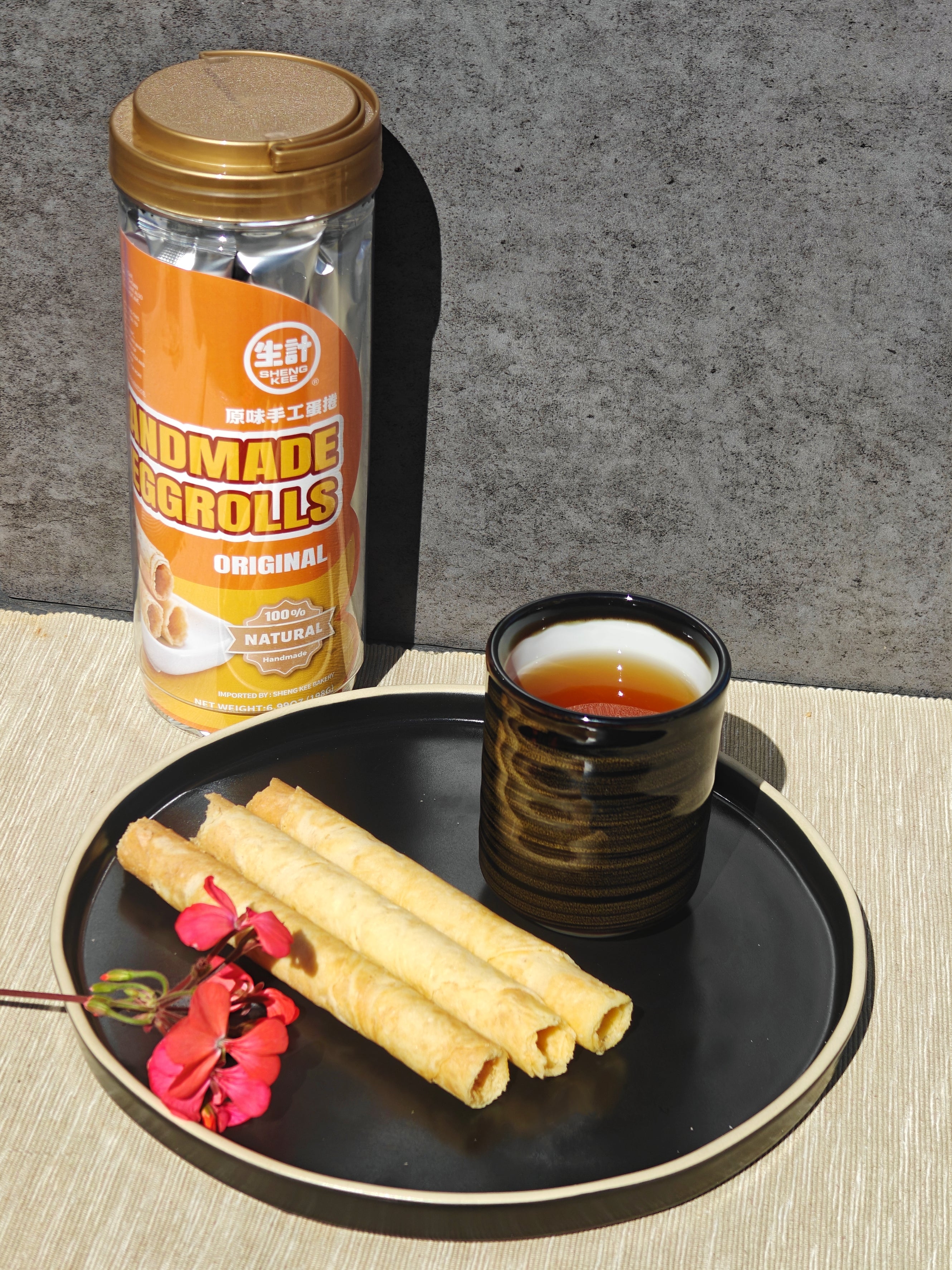 A tin of handmade egg rolls stands behind a black plate holding three rolled pastry pieces and a small cup of tea, set on a light textured surface against a gray wall.