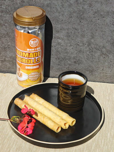 A tin of handmade egg rolls stands behind a black plate holding three rolled pastry pieces and a small cup of tea, set on a light textured surface against a gray wall.