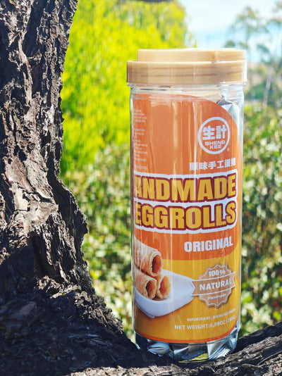 A clear plastic jar of Handmade Eggrolls with a beige lid and orange label, set on a tree trunk outdoors with a blurred green background.