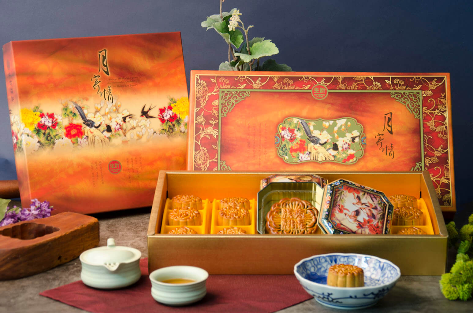 A gift box of assorted mooncakes, with the box open to display the mooncakes, a teacup, and a small round cake on a plate.
