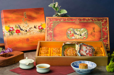 A gift box of assorted mooncakes, with the box open to display the mooncakes, a teacup, and a small round cake on a plate.