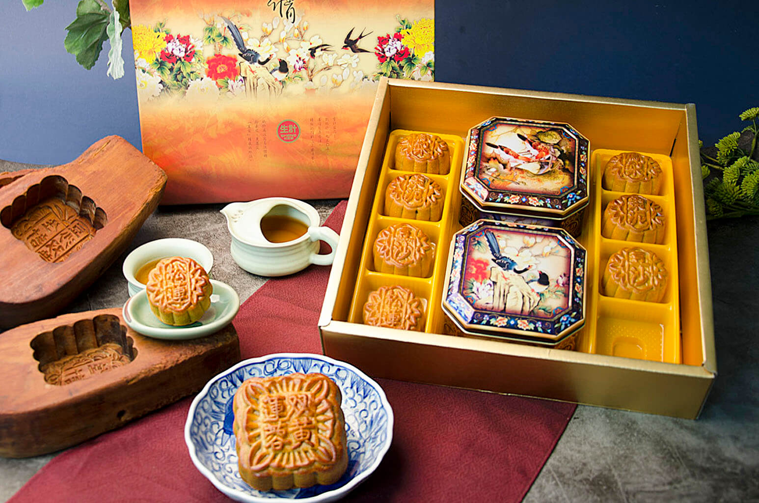 A gift box set of assorted mooncakes, with a larger mooncake displayed outside the box and eight smaller ones inside, arranged in a traditional pattern. The box is open, showing the mooncakes with decorative patterns on their tops, served alongside a cup of tea and a small plate.