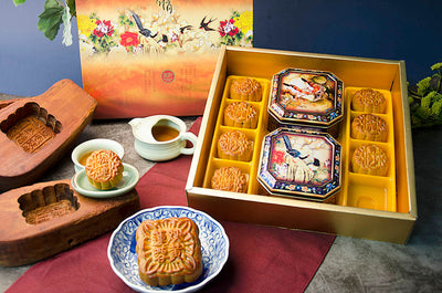 A gift box set of assorted mooncakes, with a larger mooncake displayed outside the box and eight smaller ones inside, arranged in a traditional pattern. The box is open, showing the mooncakes with decorative patterns on their tops, served alongside a cup of tea and a small plate.