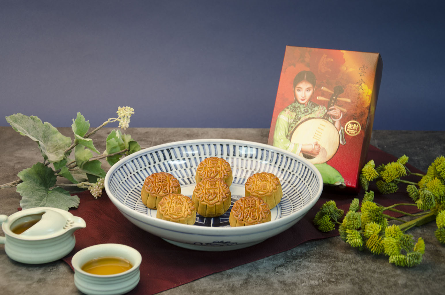 A box of assorted small mooncakes on a blue and white plate with a decorative box in the background, alongside a small cup of tea and decorative foliage.