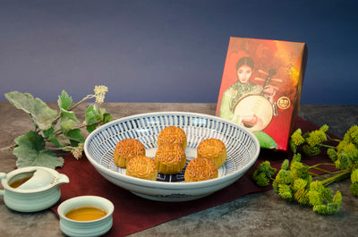 A box of assorted small mooncakes on a blue and white plate with a decorative box in the background, alongside a small cup of tea and decorative foliage.