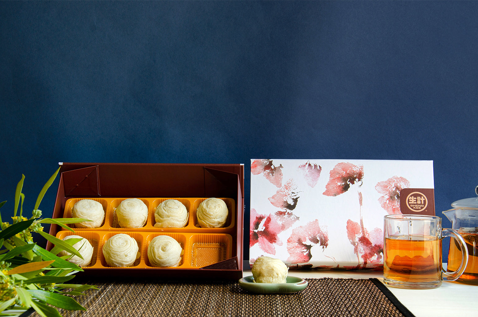 8 pcs Lava Custard Taro Pastry Box. One pastry on a small dish in front of a decorative pink, red, and white box. A glass cup of tea and tea pot beside the box.