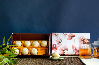 8 pcs Lava Custard Taro Pastry Box. One pastry on a small dish in front of a decorative pink, red, and white box. A glass cup of tea and tea pot beside the box.