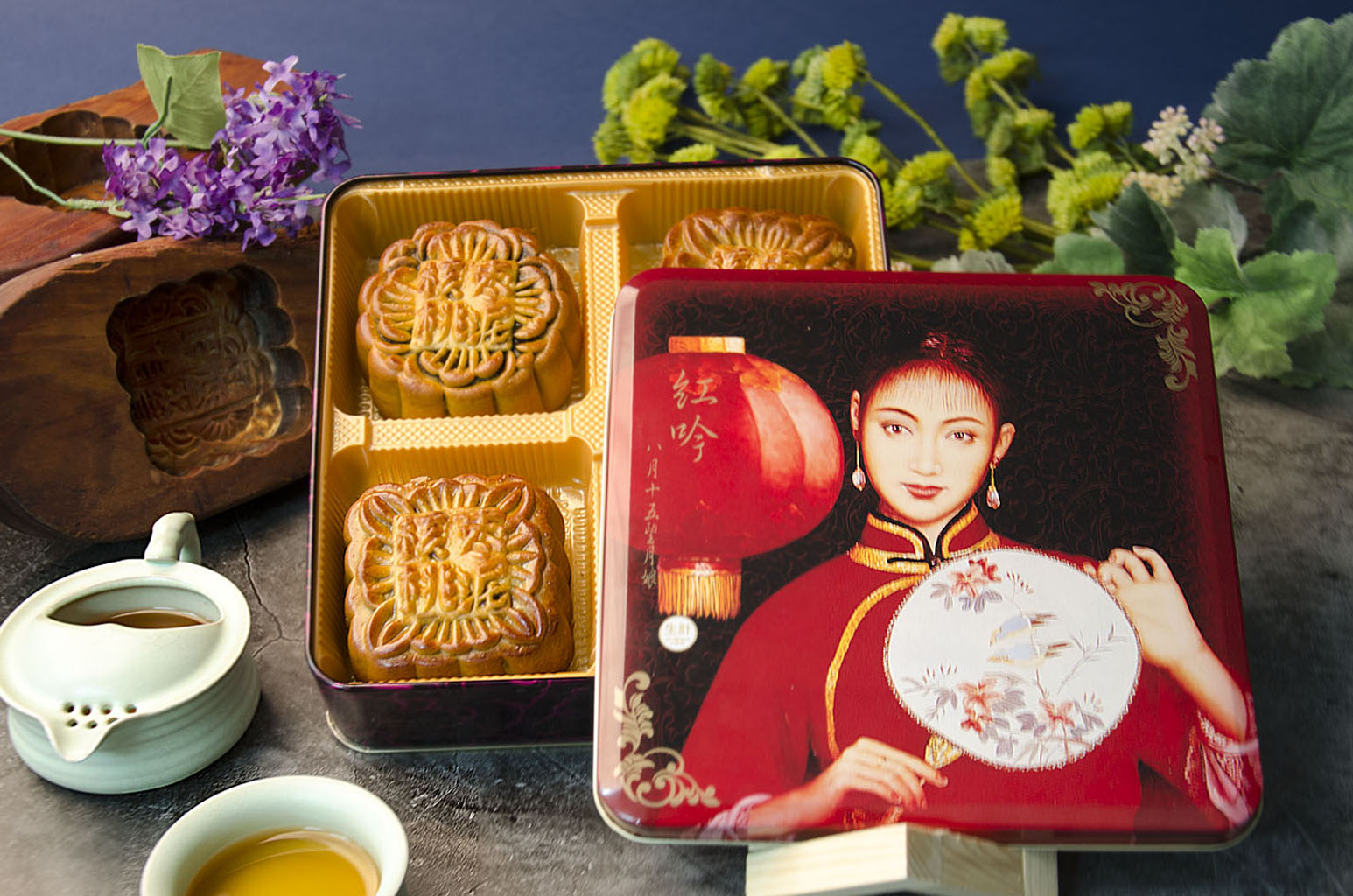 A box of Large Date Mooncakes in a decorate tin beside a cup of tea and green foliage.