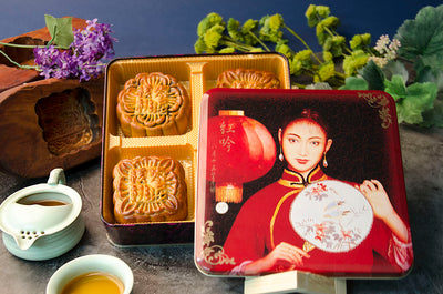 A box of Large Date Mooncakes in a decorate tin beside a cup of tea and green foliage.