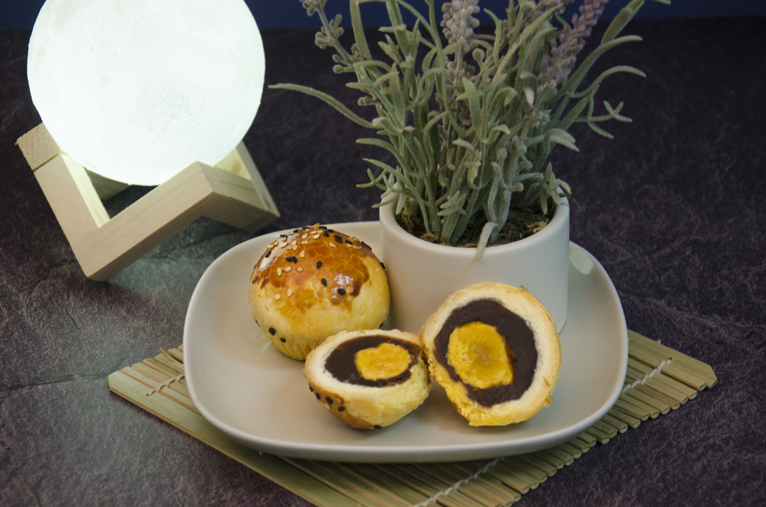 Red Bean Salted Egg Yolk Pastry. One cut open to see cross-section on a white plate with small plant in vase also on plate. In front of white ball of light on wooden contraption.