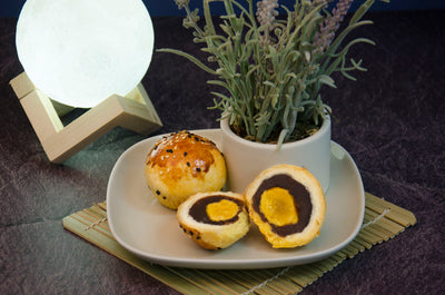Red Bean Salted Egg Yolk Pastry. One cut open to see cross-section on a white plate with small plant in vase also on plate. In front of white ball of light on wooden contraption.