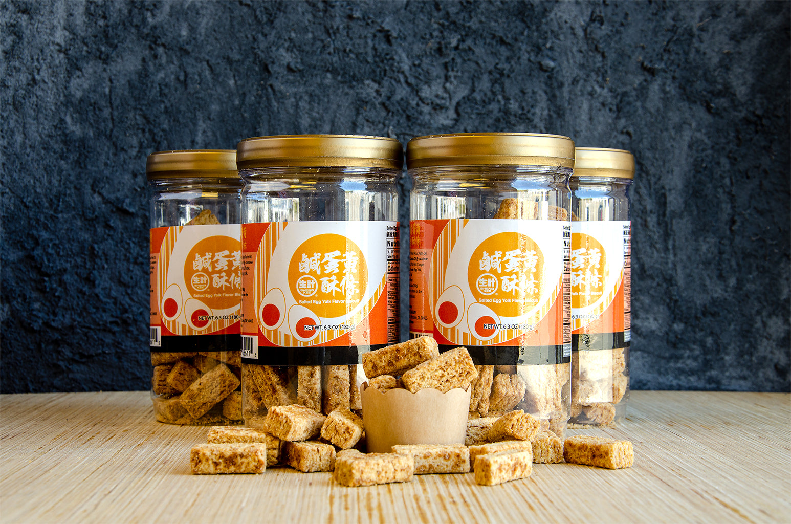 4 jars of Salted Egg Yolk Flavored Biscuits. One small cup of Salted Egg Yolk Flavored Biscuits in front of closed jars. Some Salted Egg Yolk Flavored Biscuits spilled in front of the jars and small cup.