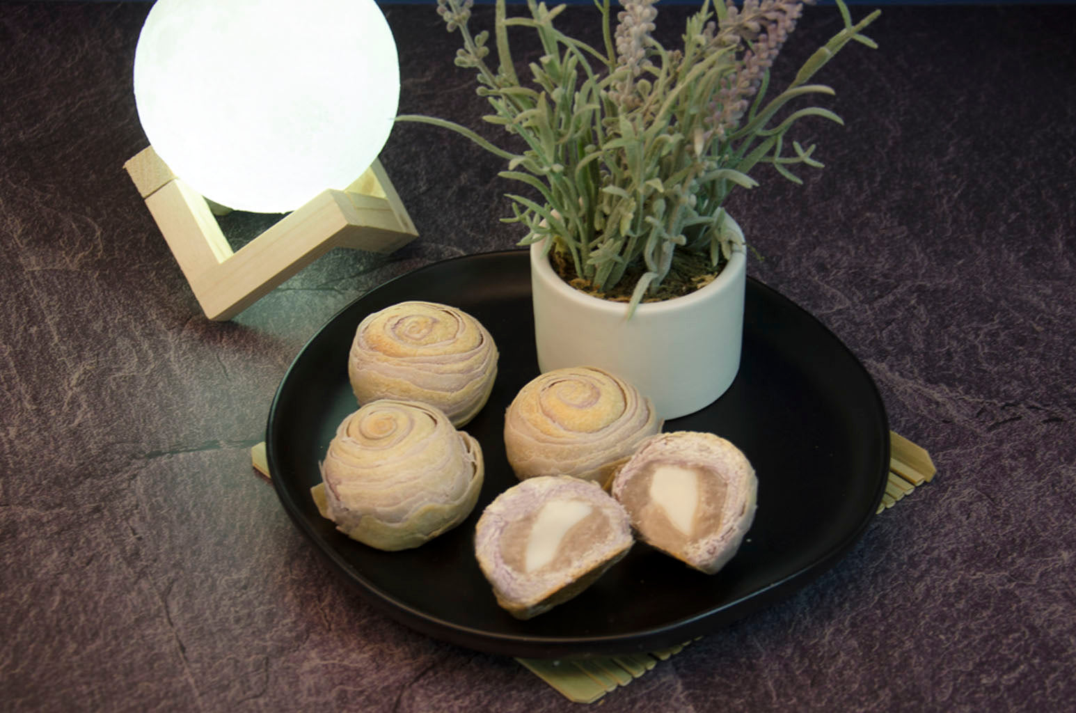 Taro Mochi Pastry with one cut open to see cross-section on black plate with small plant in vase also on plate. In front of white ball of light on wooden contraption.